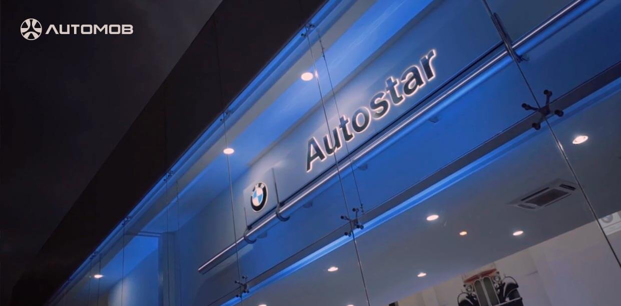  imagem retrata a fachada envidraçada e transparente de uma concessionária Autostar da marca BMW. O teto da loja, capturado durante a noite, possui iluminação azul sobre o nome da Autostar e a logomarca da BMW.
