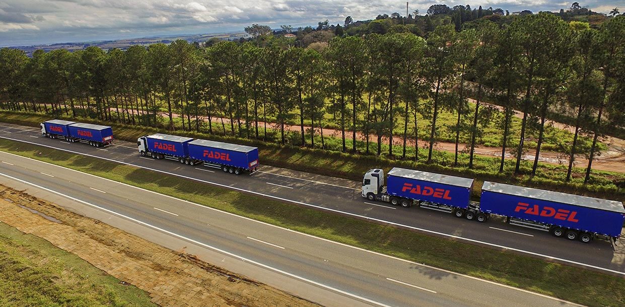 Três caminhões biarticulados, com cabines brancas e containers azuis com o logotipo da Fadel, estão numa pista de estrada durante o dia.
