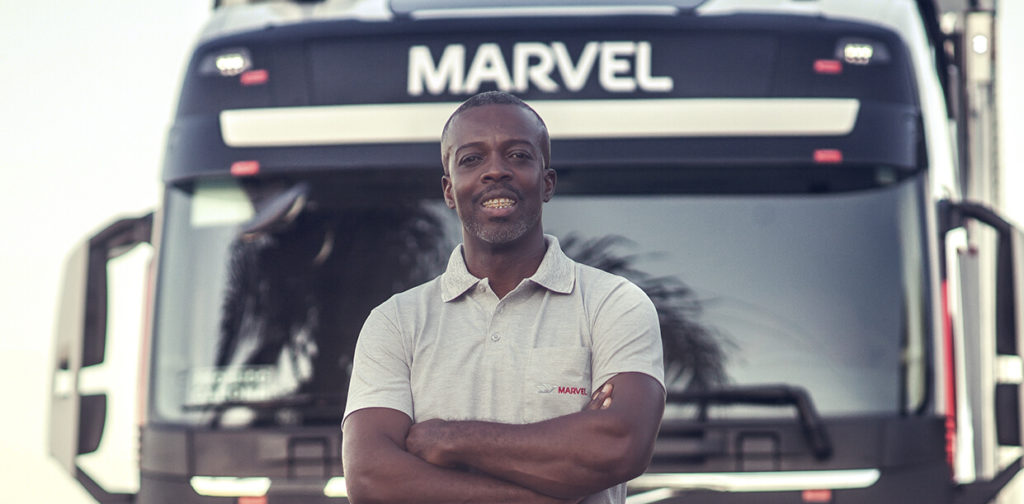 Homem com camisa polo da empresa de transportes Marvel está de braços cruzados e sorrindo à frente de um caminhão parado da mesma empresa.
