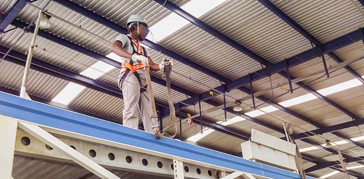 Homem com vestimentas de segurança e usando EPIs (capacete, cinto e faixas reluzentes) está em pé no alto de uma estrutura de ferro, segurando-se nas grades de proteção.
