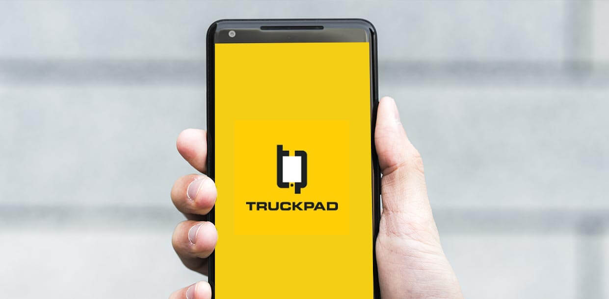 Foto mostra um celular sendo segurado pela mão de um homem. A tela do celular está mostrando o logotipo da Truckpad no centro de uma tela amarela.