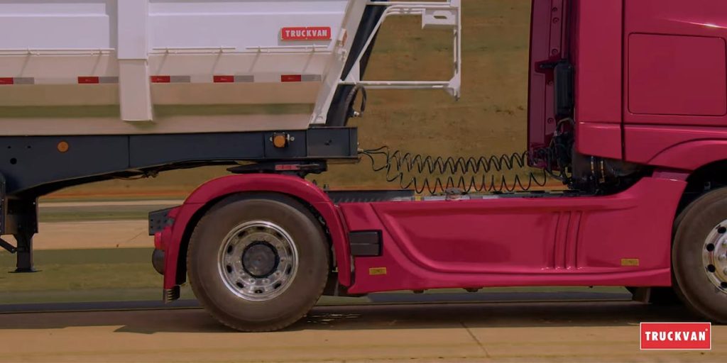 A imagem mostra a parte lateral de um caminhão vermelho transportando carga na estrada durante o dia. O logotipo da Truckvan está na parte superior do compartimento de carga.