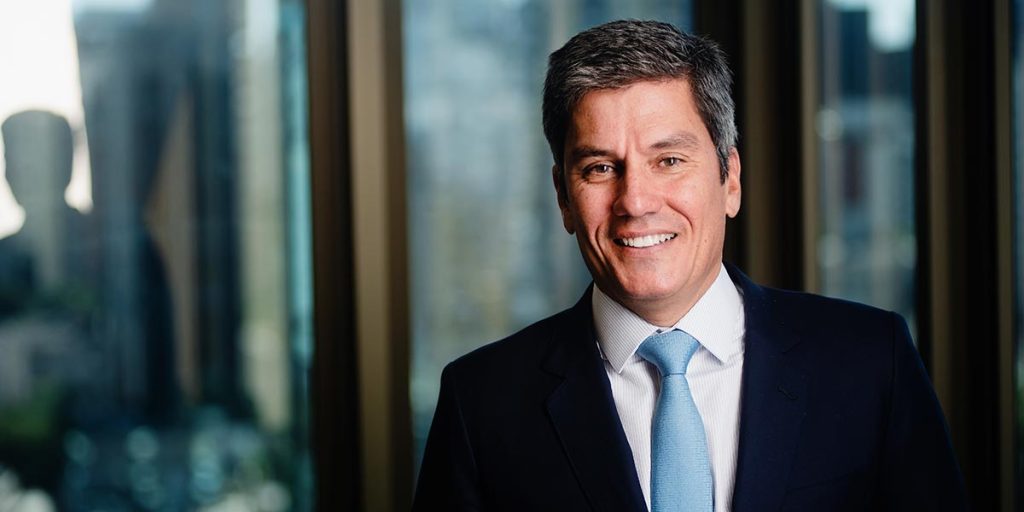 A imagem retrata o rosto sorridente de Denys Marc Ferrez, CFO do Grupo SIMPAR; homem branco de cabelos grisalhos, usando terno preto, camisa branca e gravata azul, à frente de uma janela espelhada.