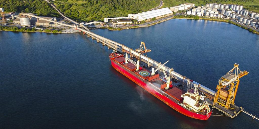 Imagem retrata uma zona portuária industrial, com silos e galpões ao longo da costa e um navio de carga vermelho aportado no mar.