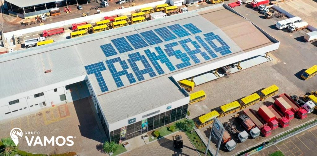 Foto tirada de cima de uma concessionária Transrio, empresa do Grupo Vamos. No telhado, painéis de energia solar escrevem Transrio, enquanto na garagem há diferentes caminhões e ônibus.