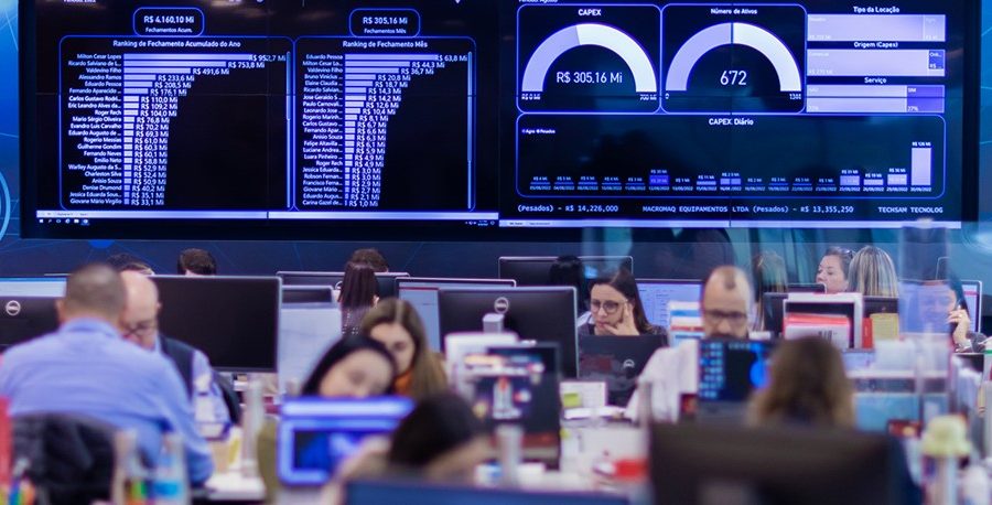 : Imagem com diversas pessoas sentadas em frente aos seus computadores trabalhando com day trade. Ao fundo e atrás delas, está um painel digital grande com índices e valores da carteira de ações.