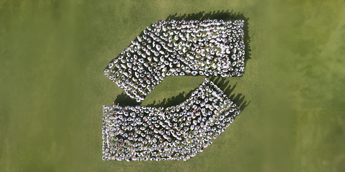 Foto com visão aérea de um campo gramado, onde pessoas estão paradas próximas umas das outras, formando o logotipo da SIMPAR.